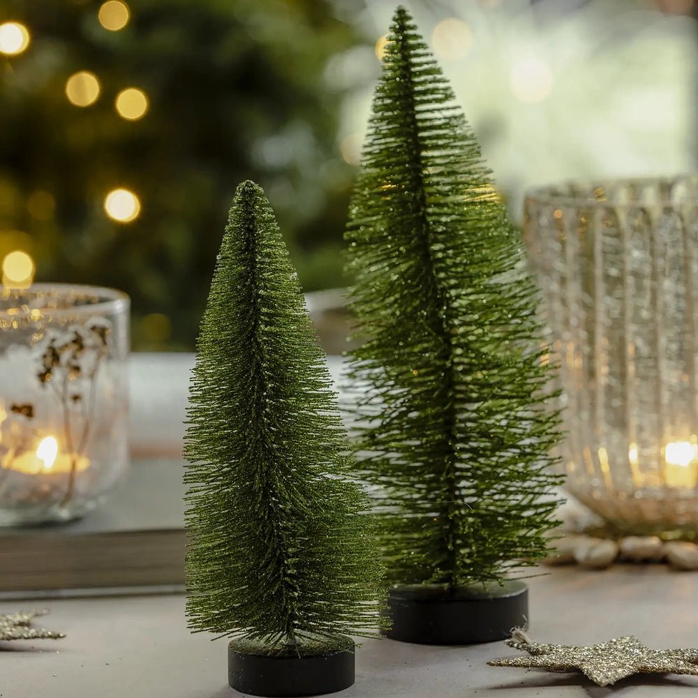 Orian large green tree stands behind smaller bottlebrush trees, showing its dense green bristles and dark round base beside a patterned candleholder and glass lantern.