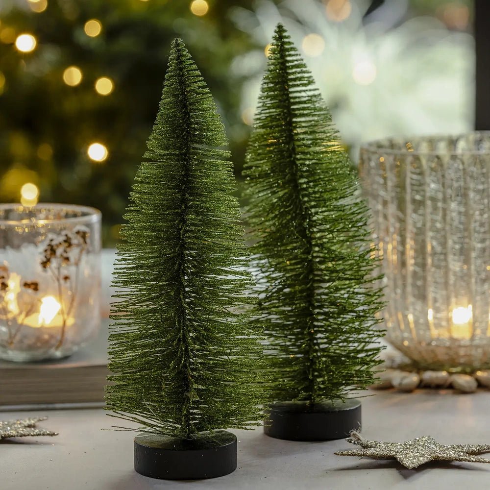 Orian large green tree displays a tall tapered bottlebrush silhouette on a dark round base, arranged beside smaller trees and glowing candleholders on a festive table.