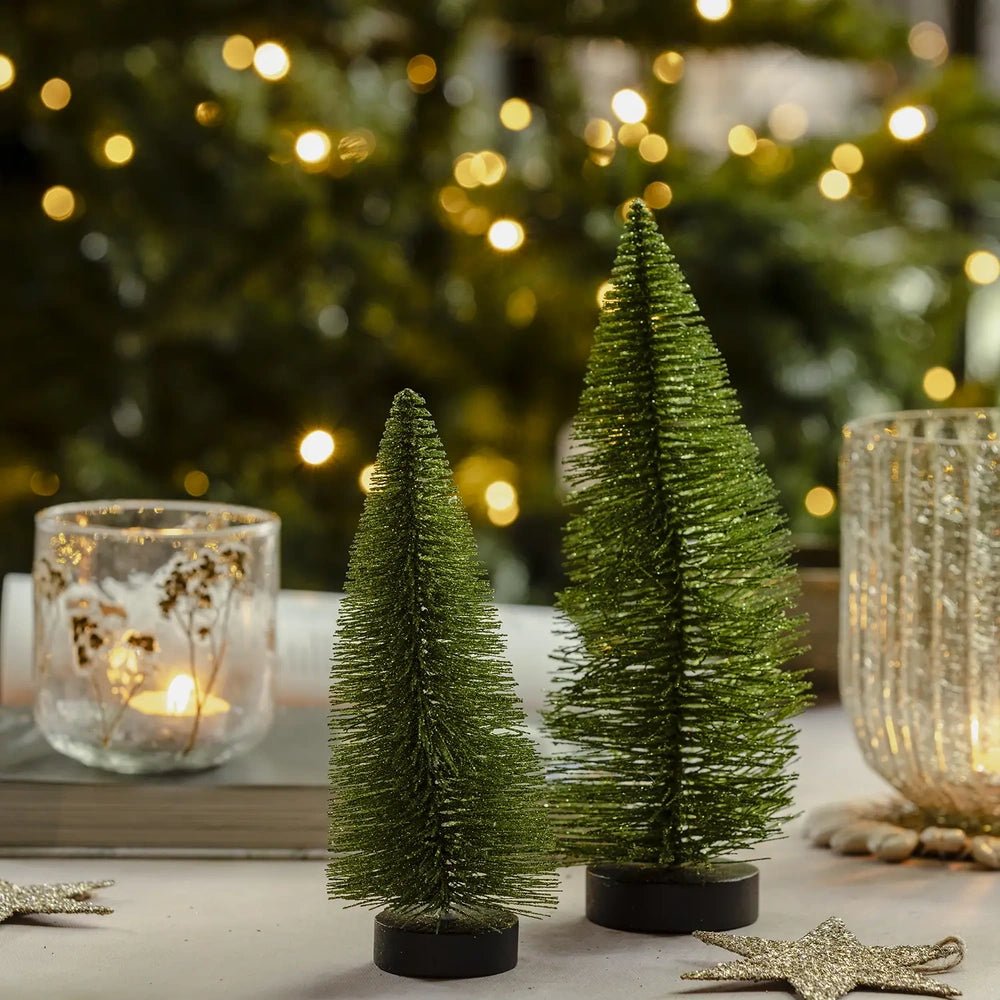 Orian large green tree forms the tallest silhouette in a trio of green bottlebrush trees on dark bases, arranged beside candleholders against warm Christmas tree lights.