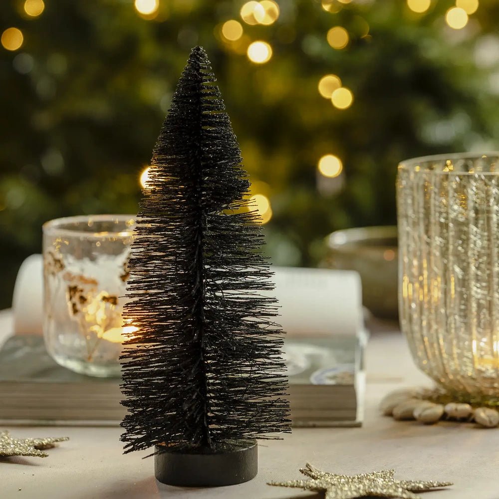 Orian large midnight tree has a dark tapered bottlebrush silhouette on a round base, displayed beside textured candleholders against softly glowing Christmas tree lights.