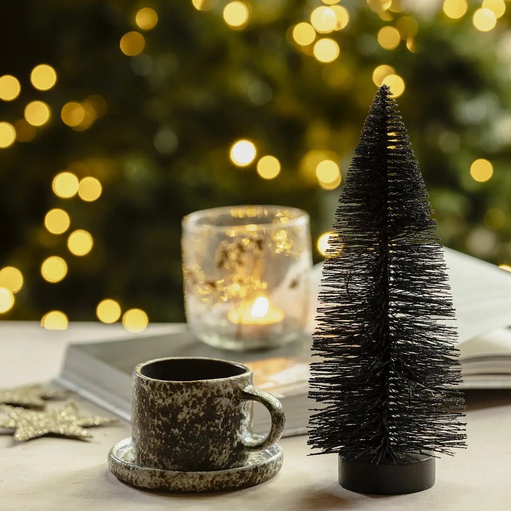 Orian medium midnight tree has a dark pointed silhouette and dense bottlebrush texture, standing beside a cup and saucer on a softly lit festive table.