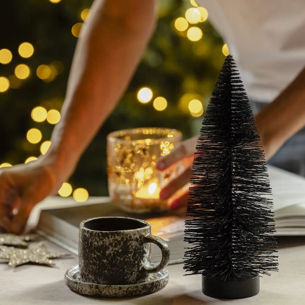Orian medium midnight tree stands beside a mottled cup and saucer while a hand arranges the nearby candleholder, with warm Christmas lights in the background.