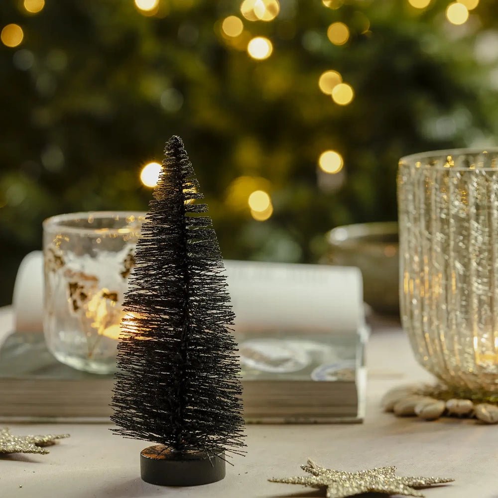 Orian medium midnight tree stands on a dark round base beside a cup and saucer, with textured candleholders and warm lights creating a festive setting.