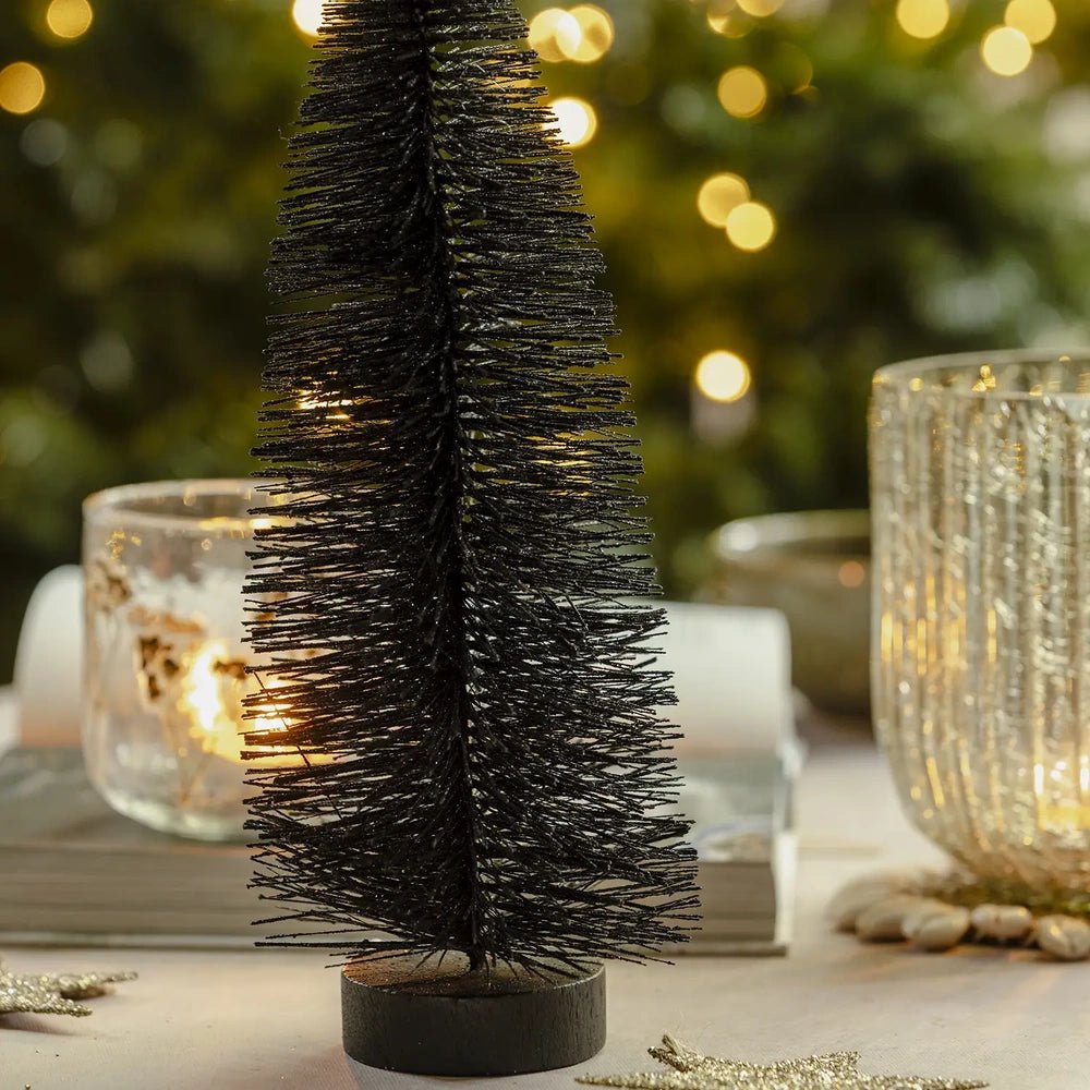 Orian midnight tree from the set of three shows dense dark bottlebrush branches and a round base, with textured candleholders and warm lights behind it.