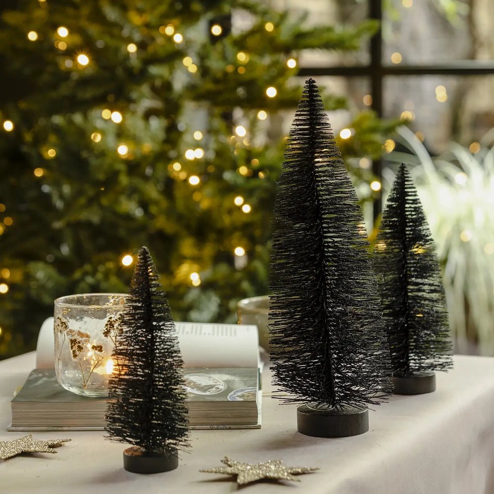 Orian midnight tree set features three graduated dark bottlebrush trees on round bases, styled with candleholders and an open book against glowing Christmas tree lights.