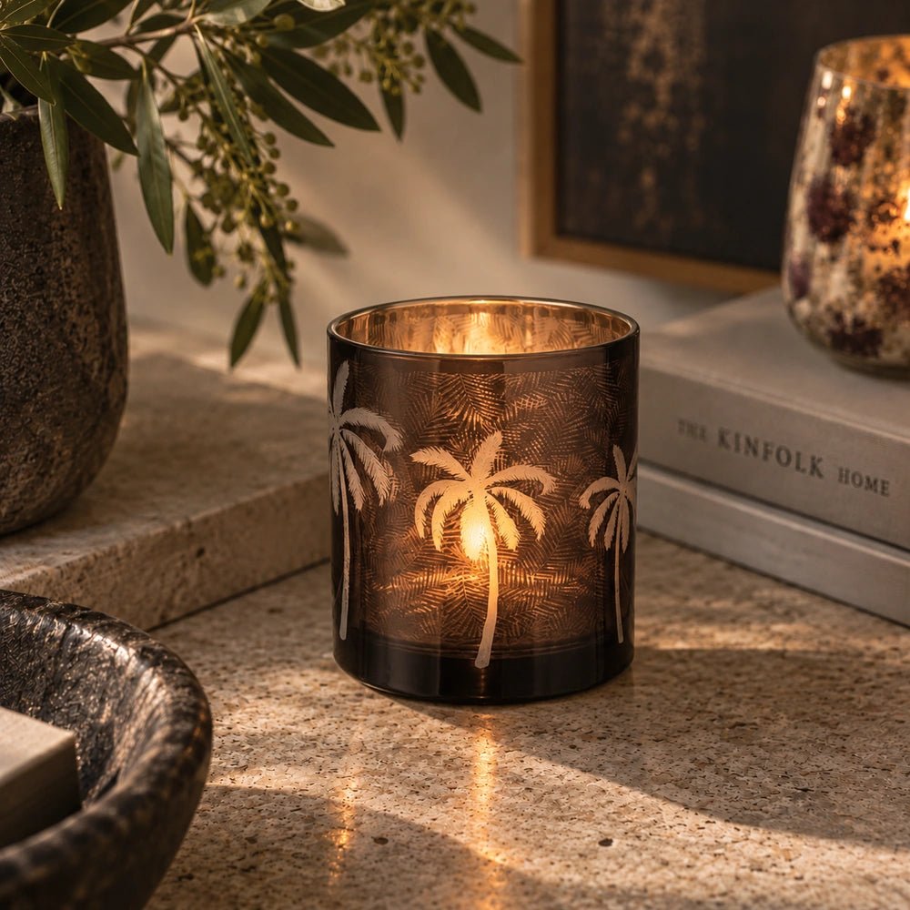 Palm Tree Glass Candle Holder glowing on a textured stone table, highlighting etched palm motifs beside greenery, stacked books and softly lit decorative glassware.