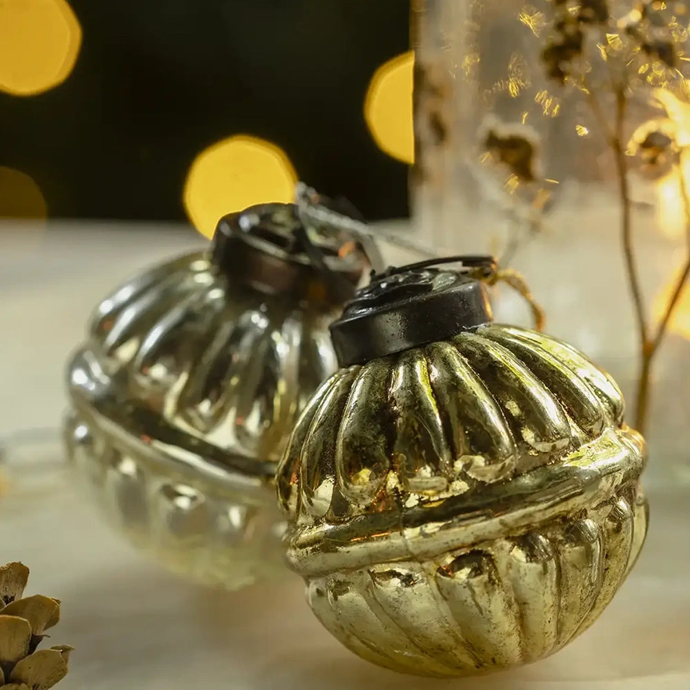 Pennington glass baubles have rounded ribbed shapes, reflective gold and silvery finishes, and dark hanging caps, resting beside a softly glowing candleholder in a festive setting.