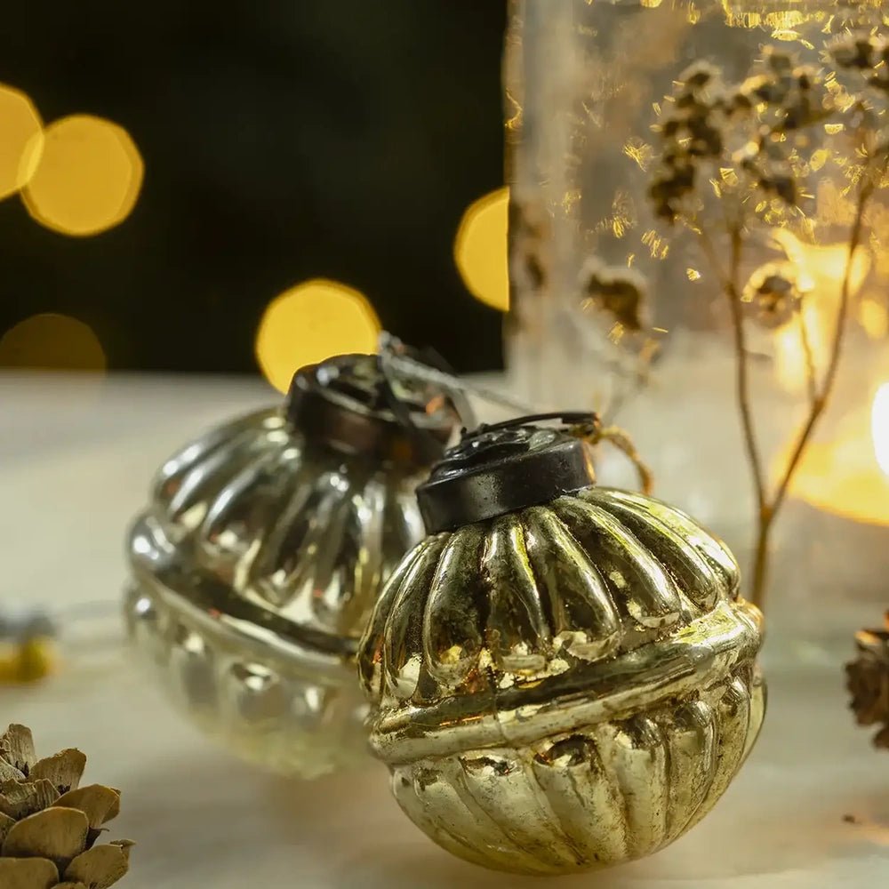Pennington glass baubles show reflective gold and silvery ribbed surfaces with dark hanging caps, styled beside a softly glowing candleholder against blurred warm Christmas lights.