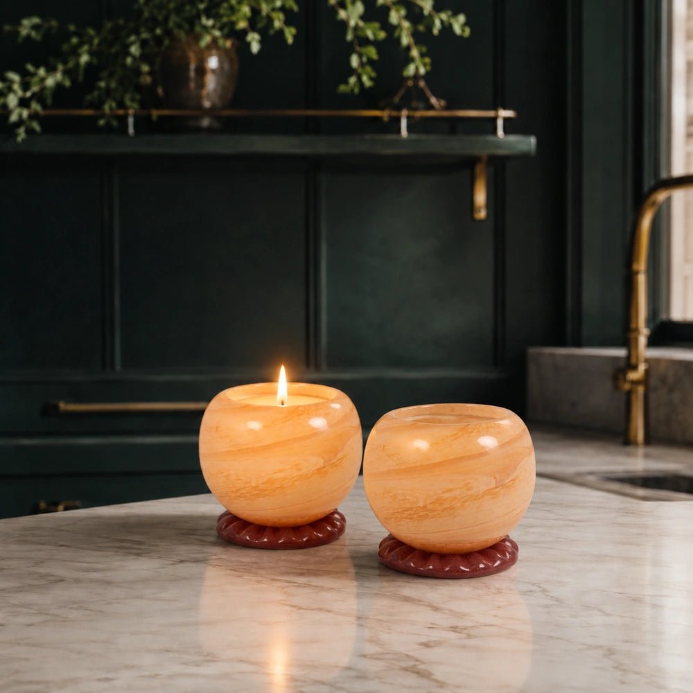 Two Ravello Glass Candle Holder, Peach candles glowing on a pale marble table, showing translucent peach glass beside dark cabinetry and softly styled foliage.