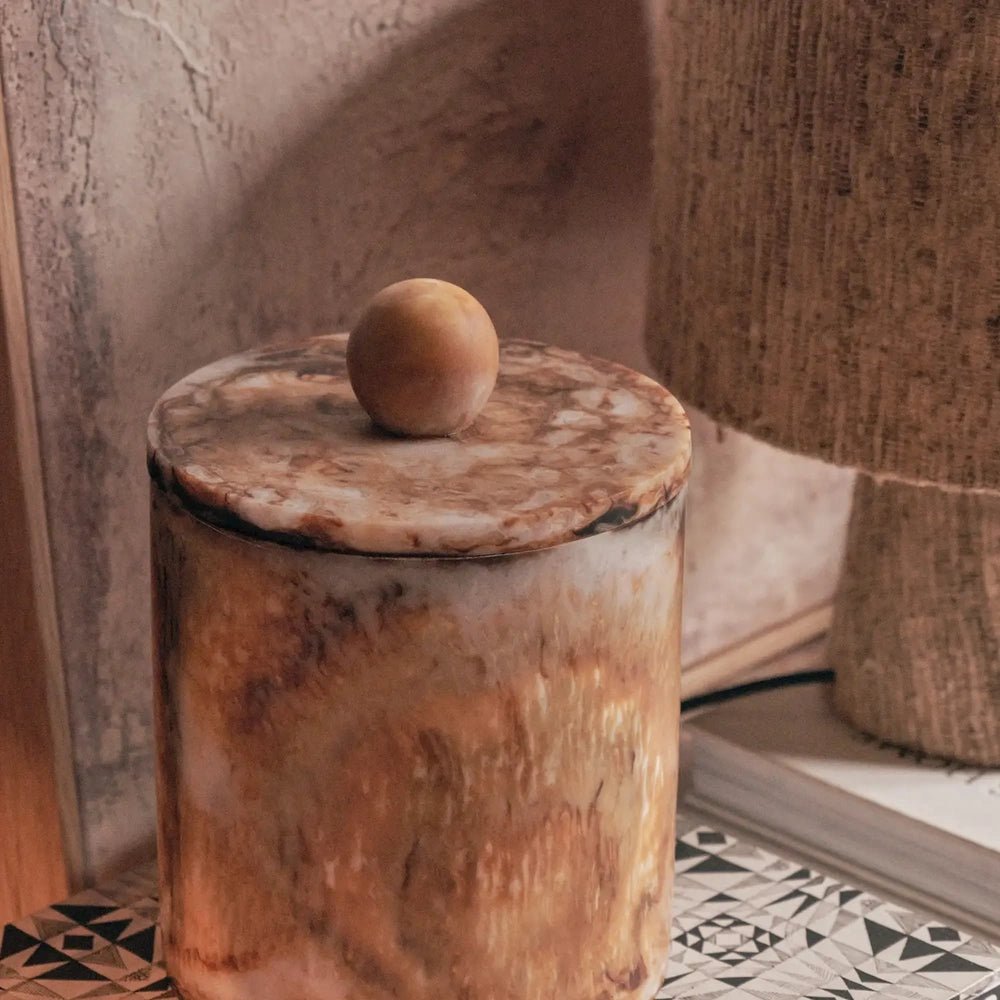 Reeve marble effect jar with an amber mottled surface and round lid knob, shown close up beside a textured lamp against a warm plaster wall.