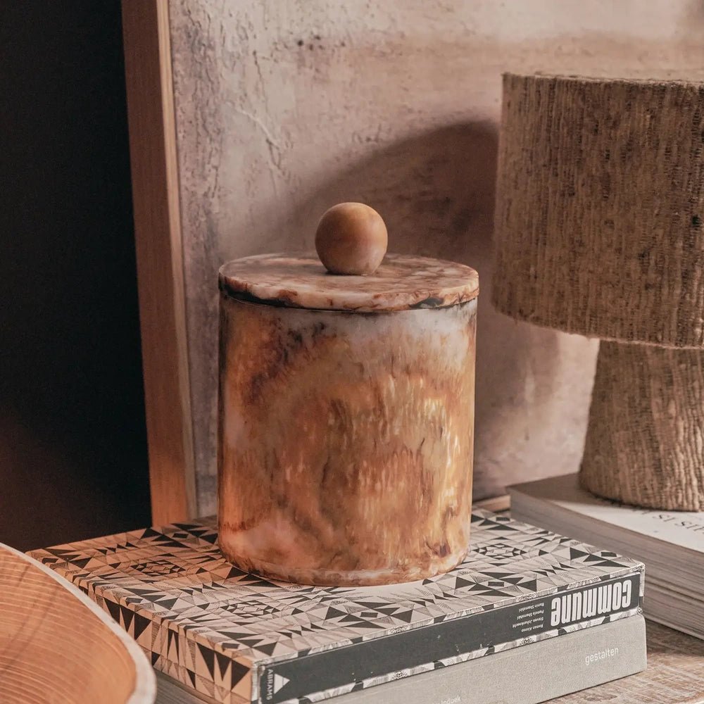 Reeve marble effect jar with a cylindrical shape, amber patterned surface and rounded lid knob, arranged on stacked books beside a neutral textured lamp.