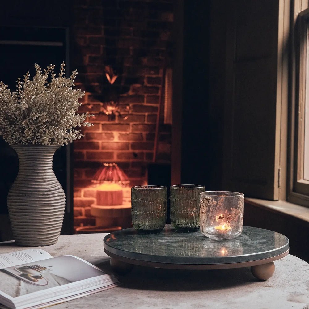 Remington decorative tray with a round green surface and rounded wooden feet, holding glasses and a candle on a coffee table beside a glowing fireplace.