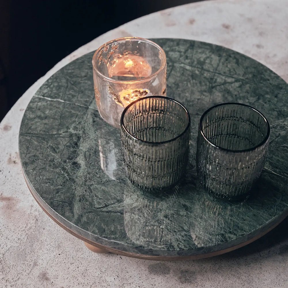 Remington decorative tray viewed from above, showing its circular green marbled surface beneath two ribbed glasses and a glowing candle on a pale stone table.