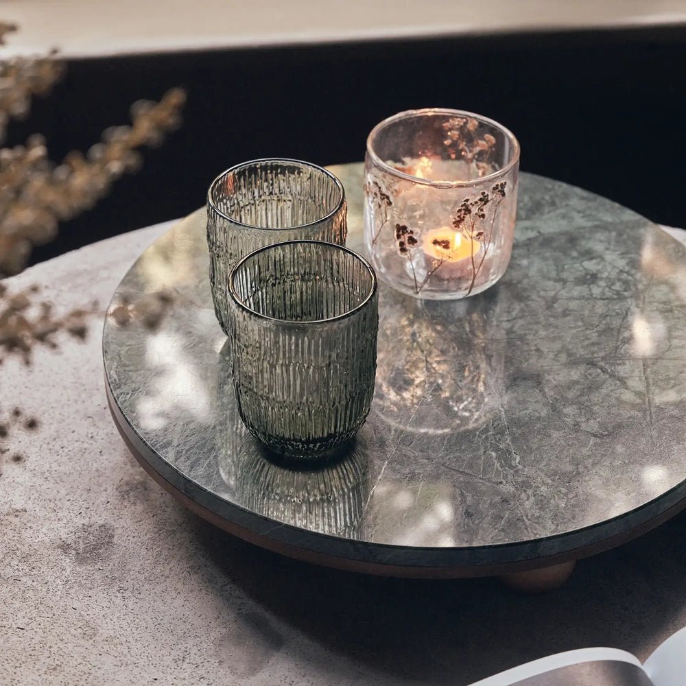Remington decorative tray with a round reflective green patterned surface, holding two ribbed glasses and a glowing candle on a pale table beside a dark wall.