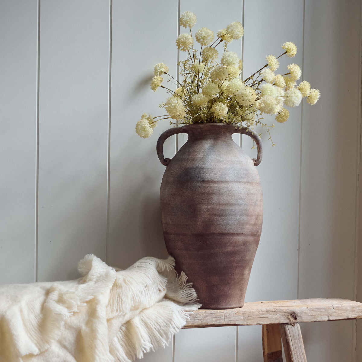 The Romy Terracotta Vase, a tall amphora shaped vase with two handles in speckled brown glaze, containing creamy meadow stems.
