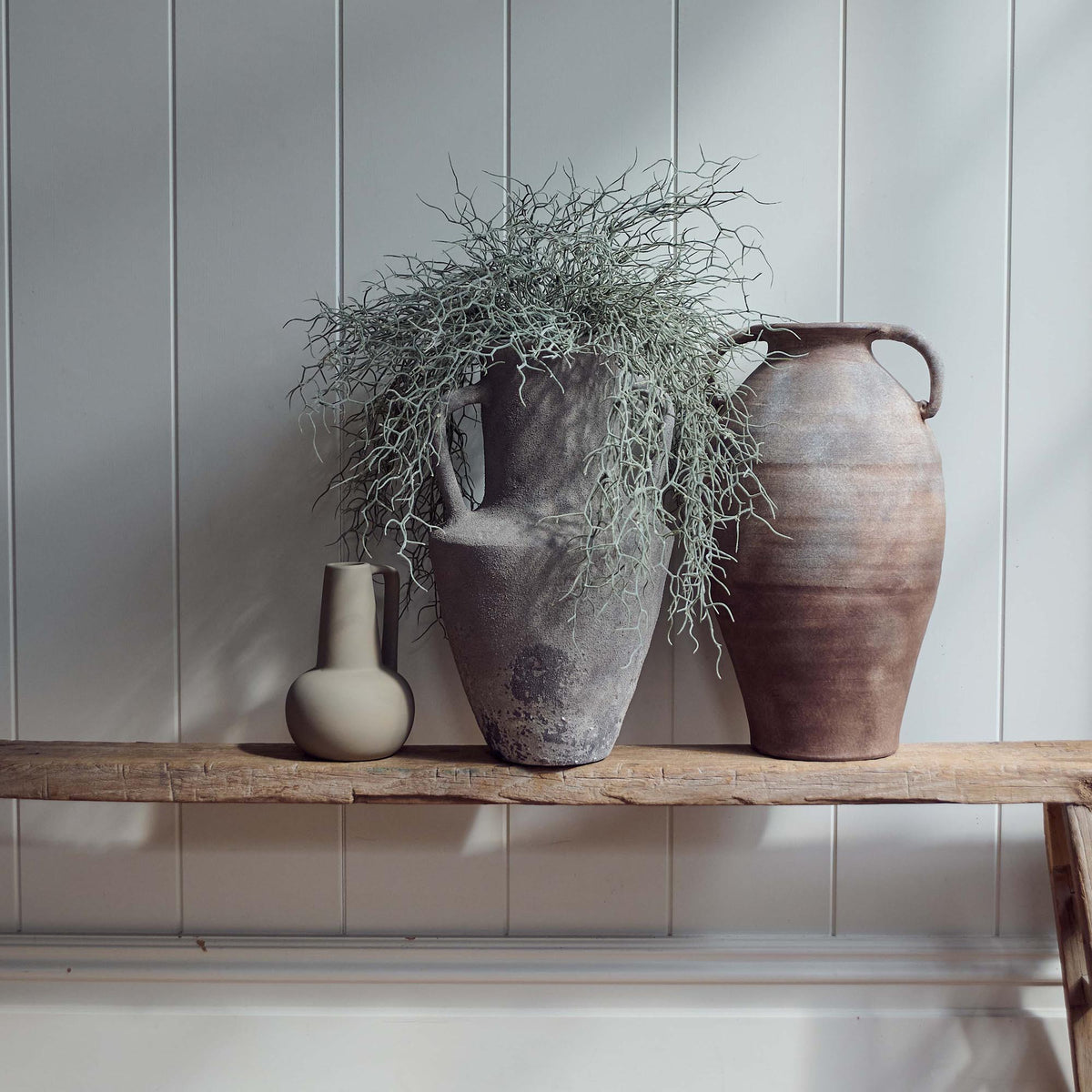 The Rowan Terracotta Vase, an angular grey amphora shaped vase with two handles holding some wispy faux trailing green stems.