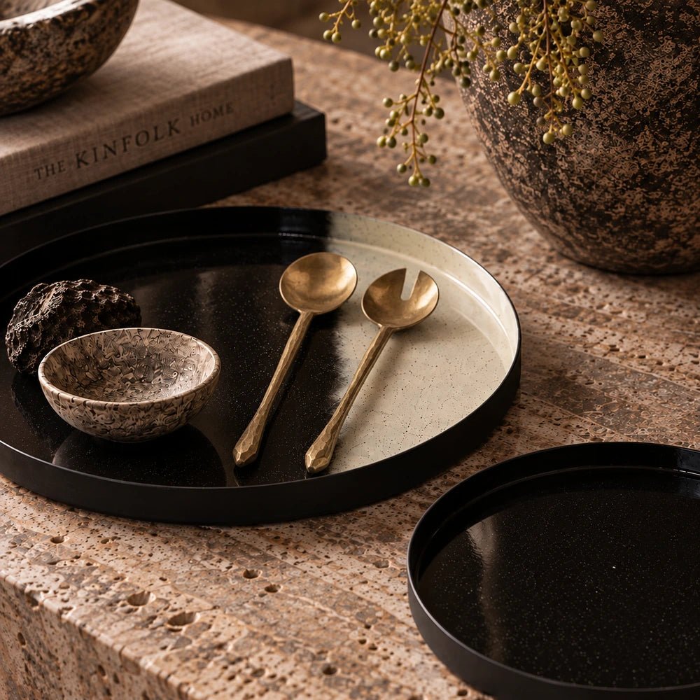 Rooke Metal Salad Servers arranged on a black tray atop a textured table, styled beside a patterned bowl, stacked books and trailing greenery.
