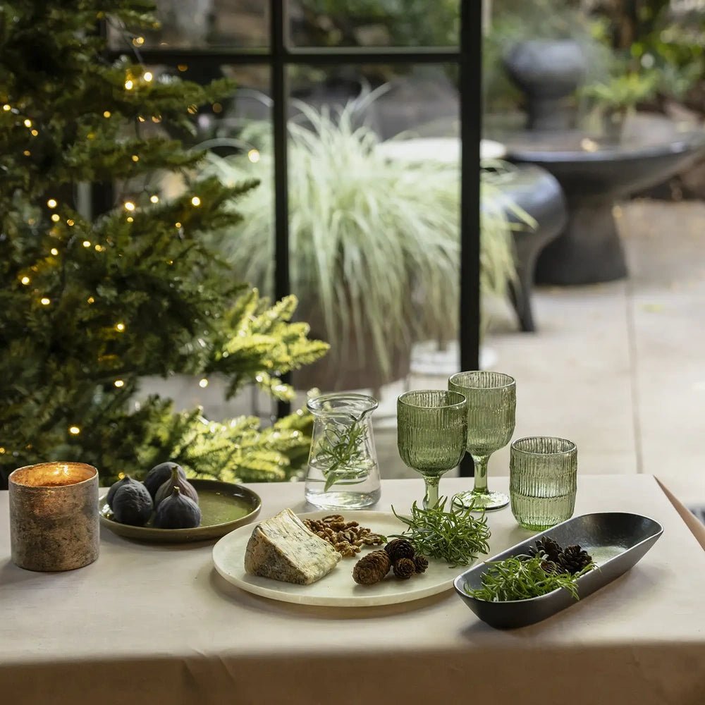 Salome round platter has a pale mottled surface, styled with cheese, walnuts and greenery beside green glasses and a candle on a festive dining table.
