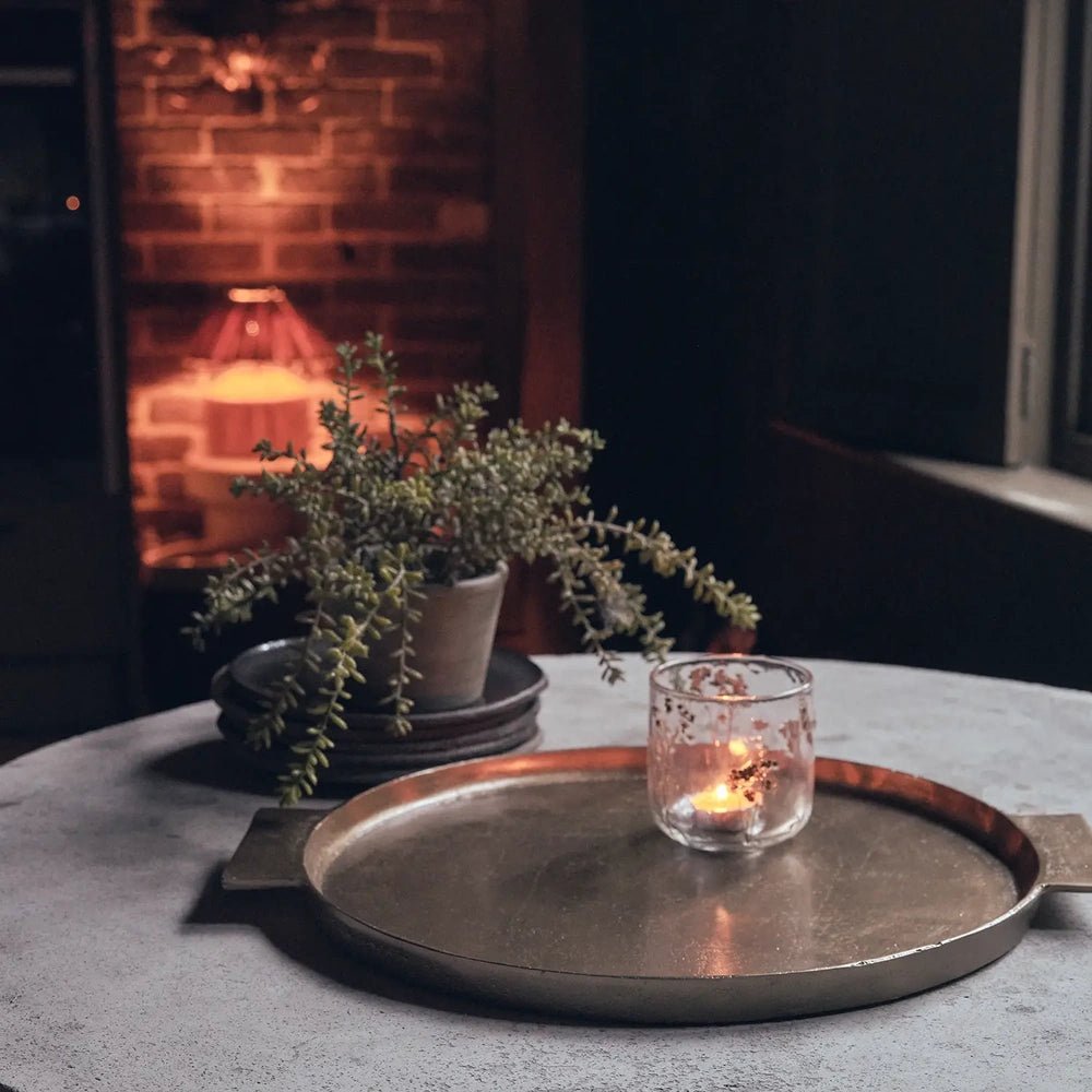 Sandford metal decorative tray with a shallow round surface and rectangular handles, holding a candle on a coffee table beside a plant and glowing fireplace.