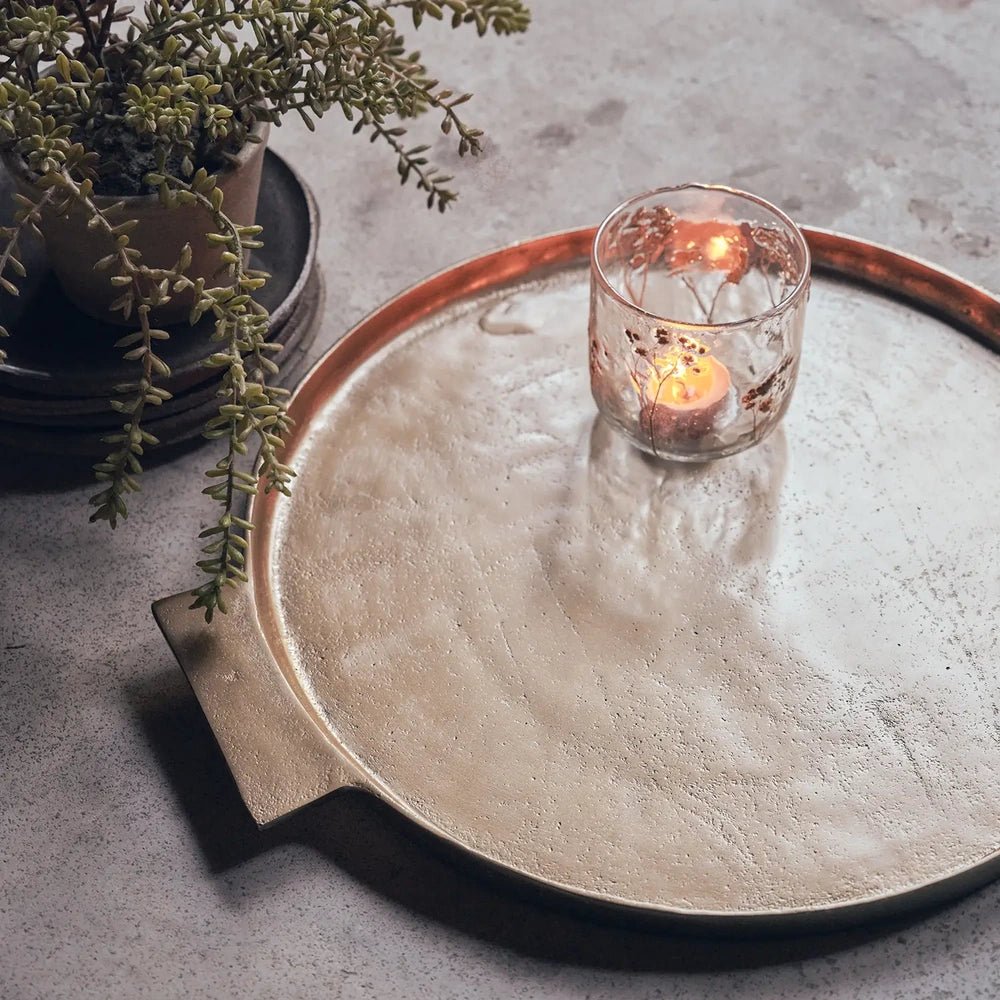 Sandford metal decorative tray viewed from above, showing its round warm toned surface and flat handles, styled with a lit candle beside a trailing plant.