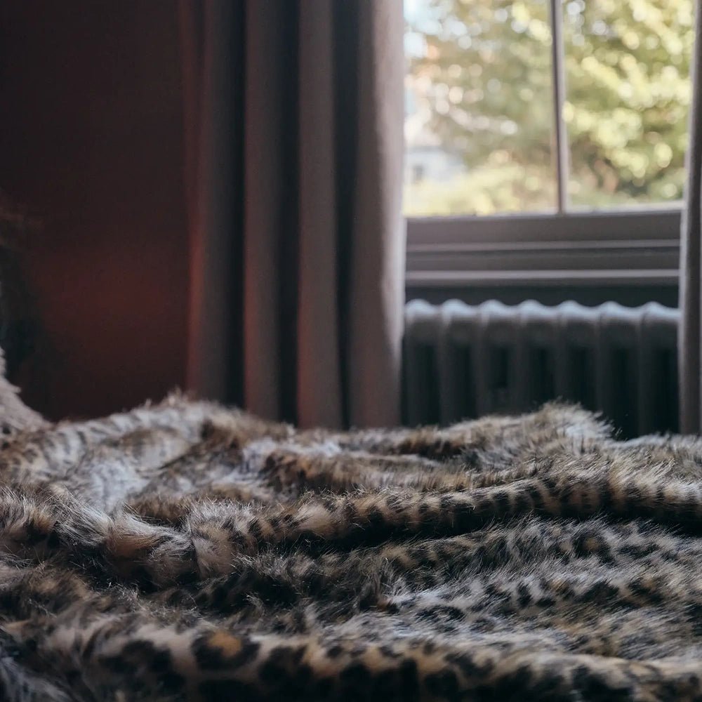 Savannah throw draped in soft folds beside a window, highlighting its dense textured pile and irregular animal print in warm brown, cream and dark tones.