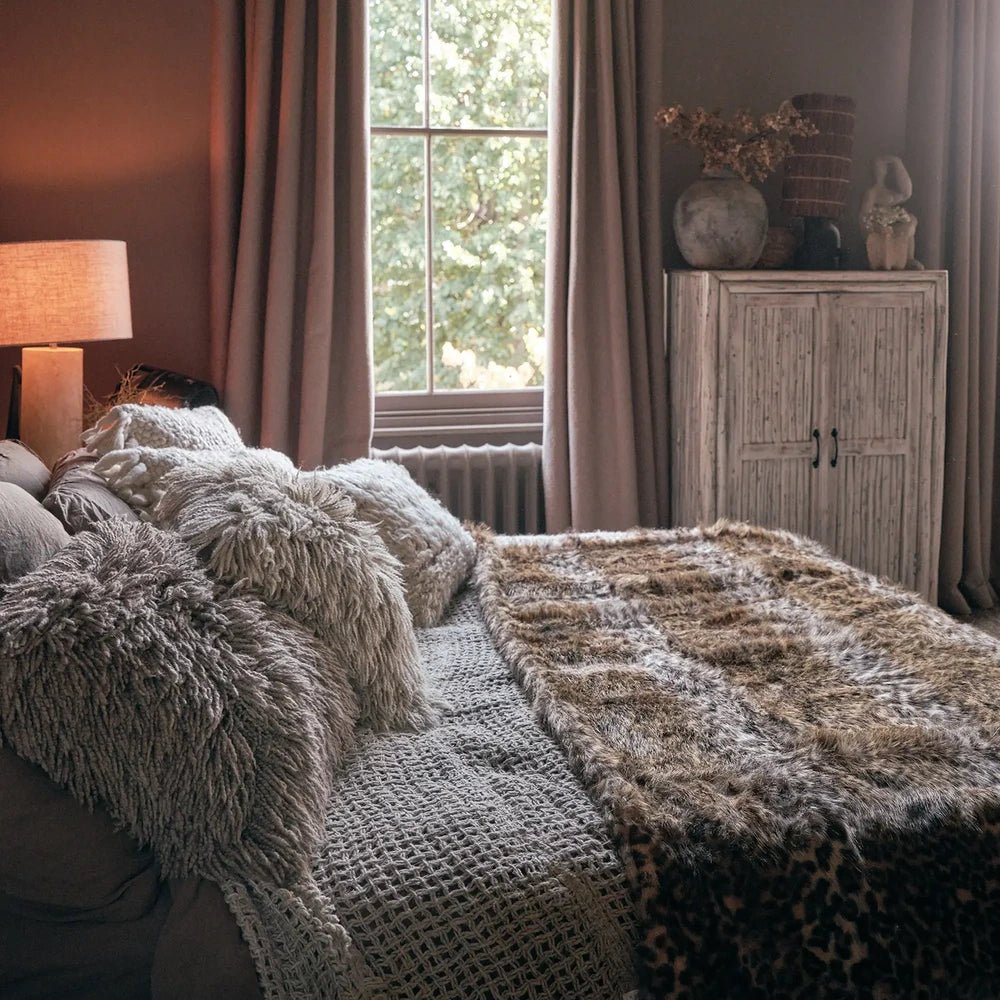 Savannah throw draped across a woven sofa with shaggy neutral cushions, showing its brown and cream pattern beside a window and pale wooden cabinet.