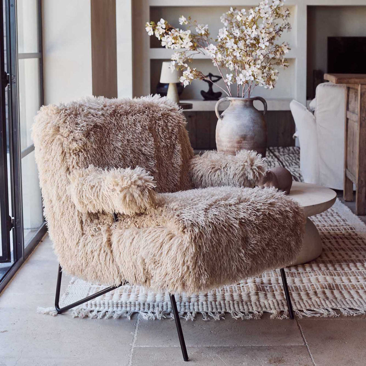 Fluffy beige armchair in a living room setting with a vase of flowers on a shelf.