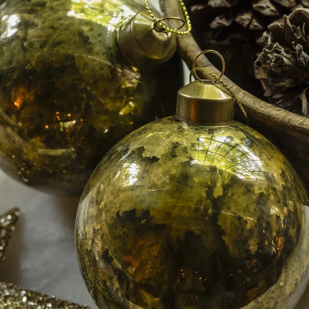 Silas antique gold bauble displays a mottled reflective finish and rounded shape, with its gold hanging cap visible beside another bauble and pine cones.
