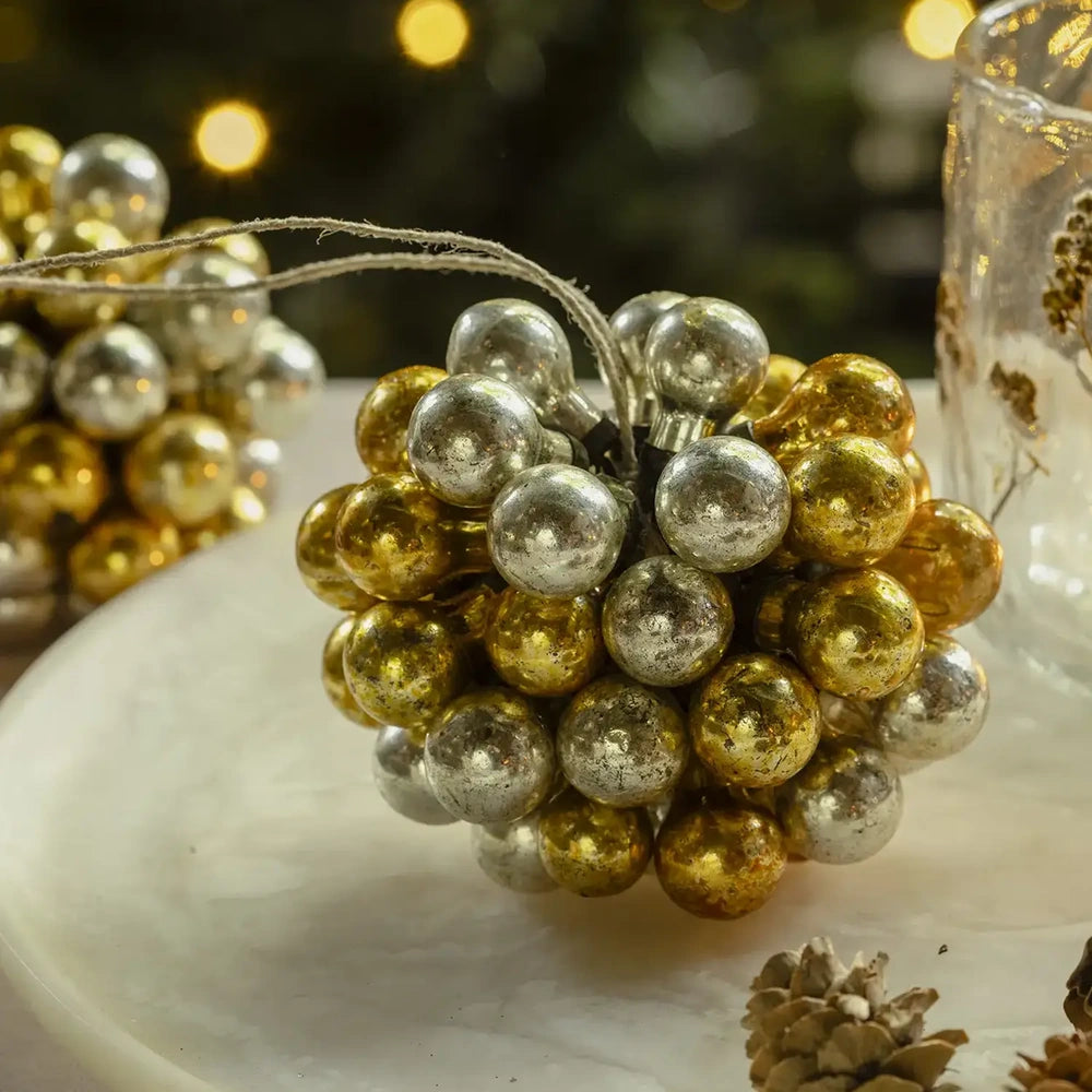 Silvero glass bauble combines clustered gold and silver spheres with a hanging cord, displayed beside a candleholder and pine cones against warm festive lights.