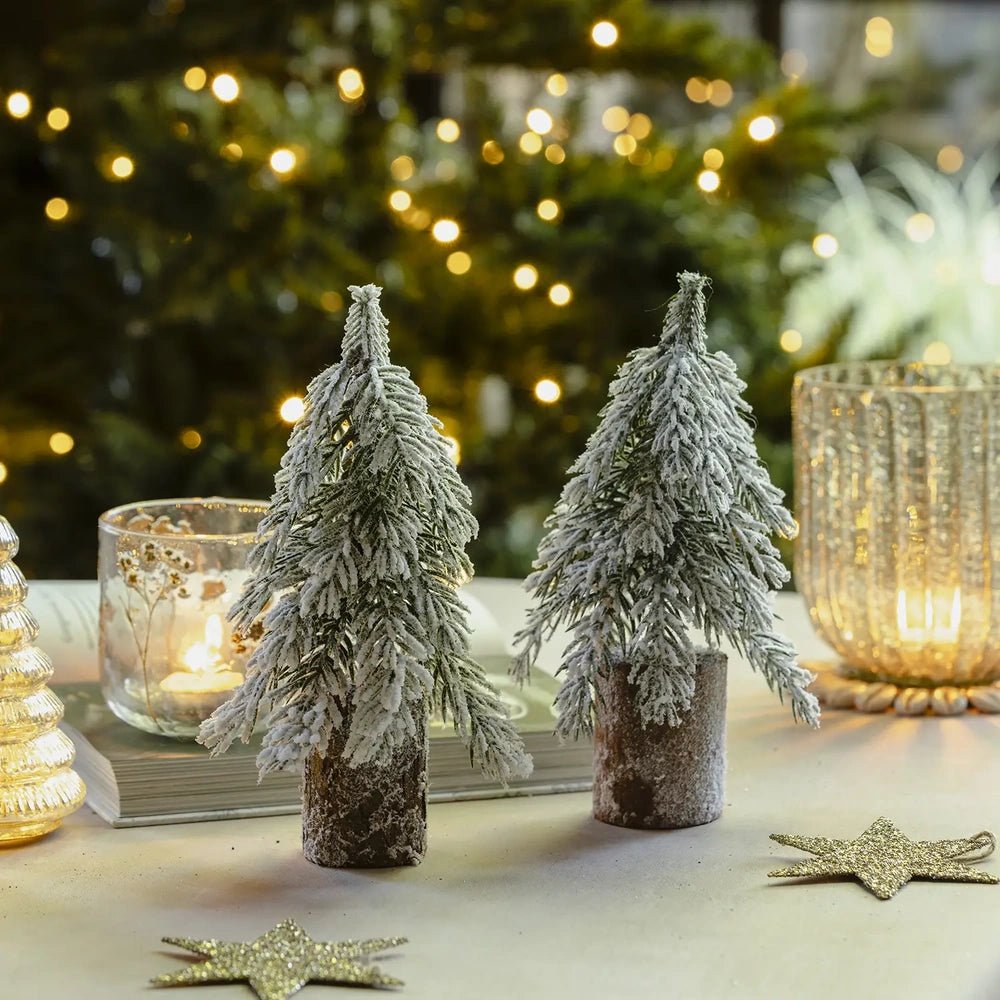 Snowy tree decorations show frosted green branches above brown cylindrical bases, styled beside glowing candleholders and a star decoration against softly blurred Christmas lights.