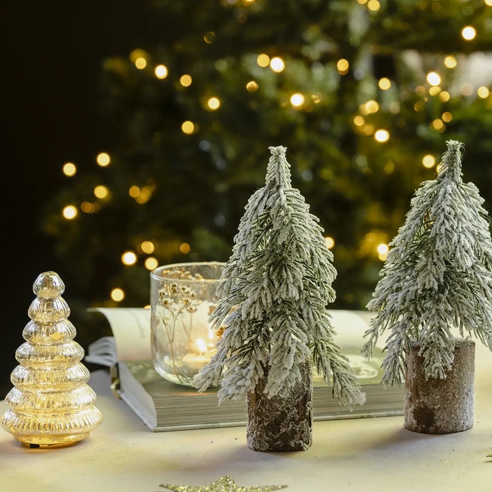 Snowy tree decorations have snow-dusted green branches and rustic brown bases, displayed as a pair beside candleholders and a small illuminated tree on a festive table.
