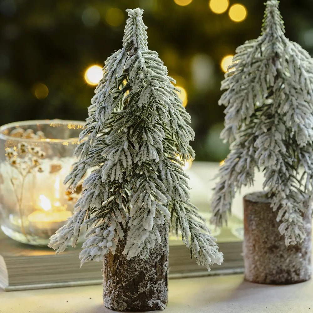 Snowy tree decoration shows thick white frosting across layered green branches and a brown cylindrical base, with a second tree and glowing candleholder behind it.