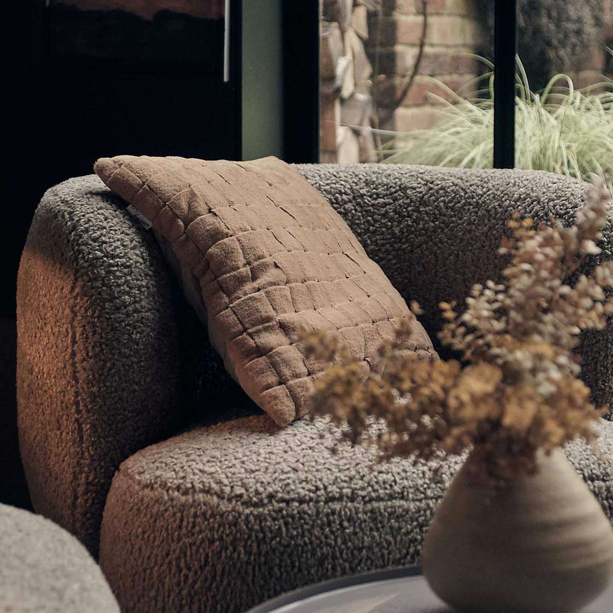 A rust coloured cushion with a checkered texture on a boucle sofa with a vase of faux flowers in front.
