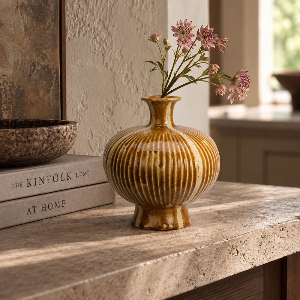 Tallis Stoneware Vase with ribbed ochre surface, styled with delicate pink flowers on a pale stone console beside stacked neutral books in warm natural light.