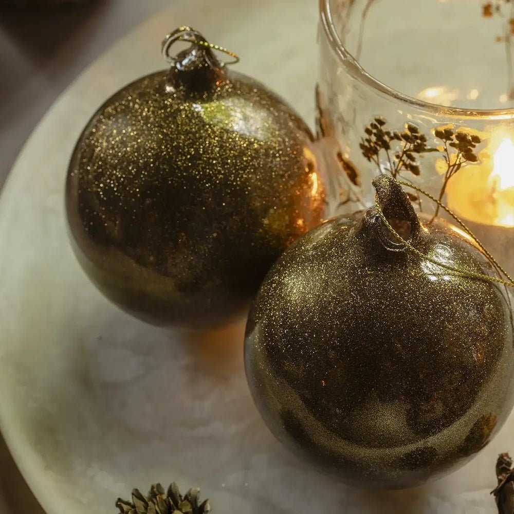 Tarim copper baubles display rounded silhouettes and finely glittered surfaces, resting on a pale platter beside pine cones and a softly glowing patterned candleholder.