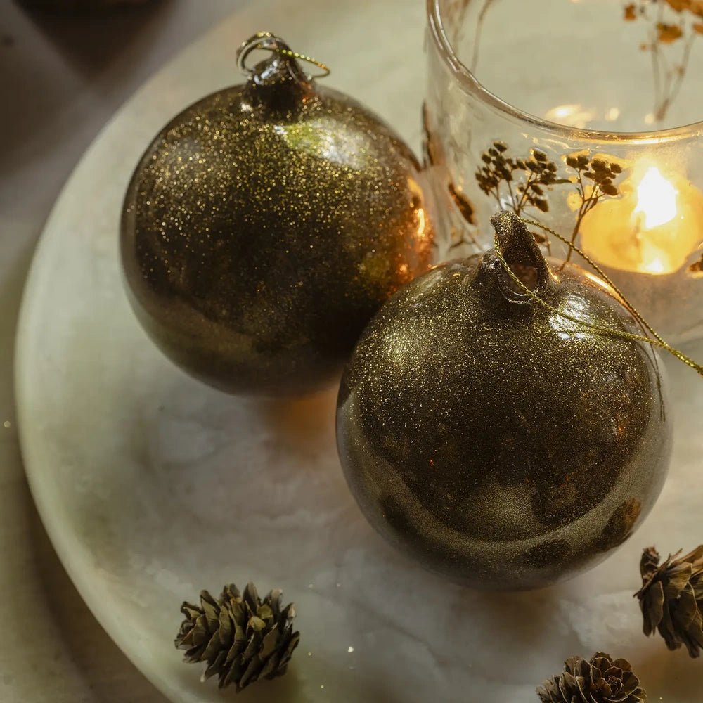 Tarim copper baubles have rounded shapes and fine glittering surfaces, displayed as a pair beside a glowing candleholder and pine cones on a pale platter.
