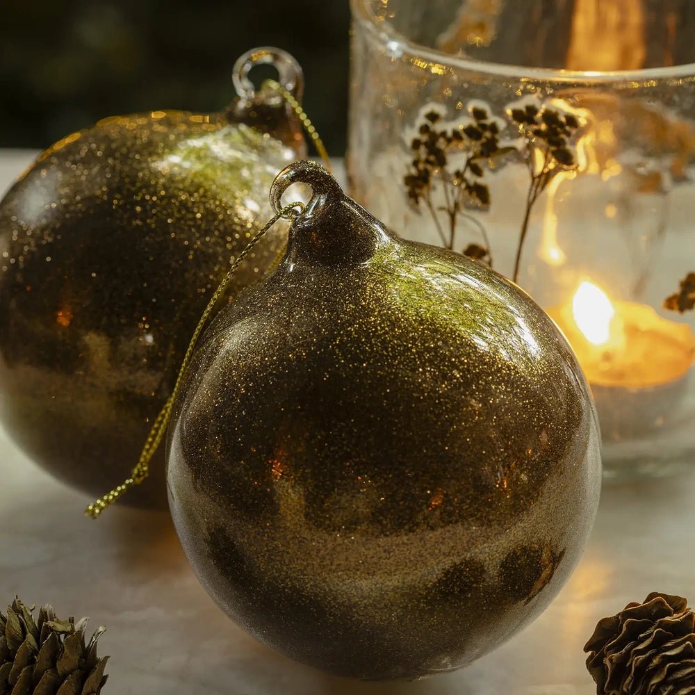 Tarim copper baubles show sparkling copper-toned surfaces with darker hanging caps, arranged beside a glowing candleholder with one rounded bauble positioned behind the other.
