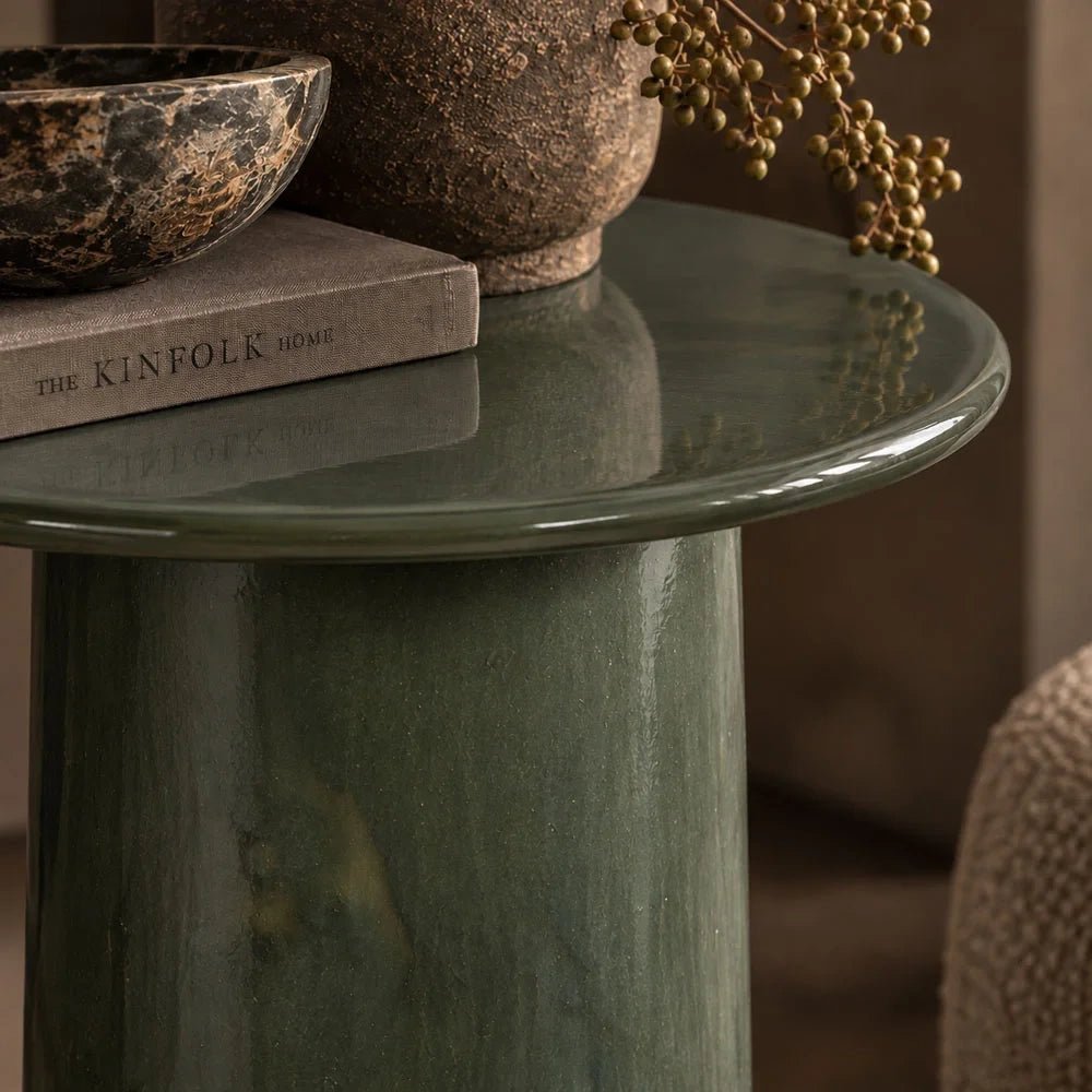 Thayer stoneware side table shown close up, highlighting its round glossy green top and cylindrical mottled base beneath a stack of books and decorative vessels.