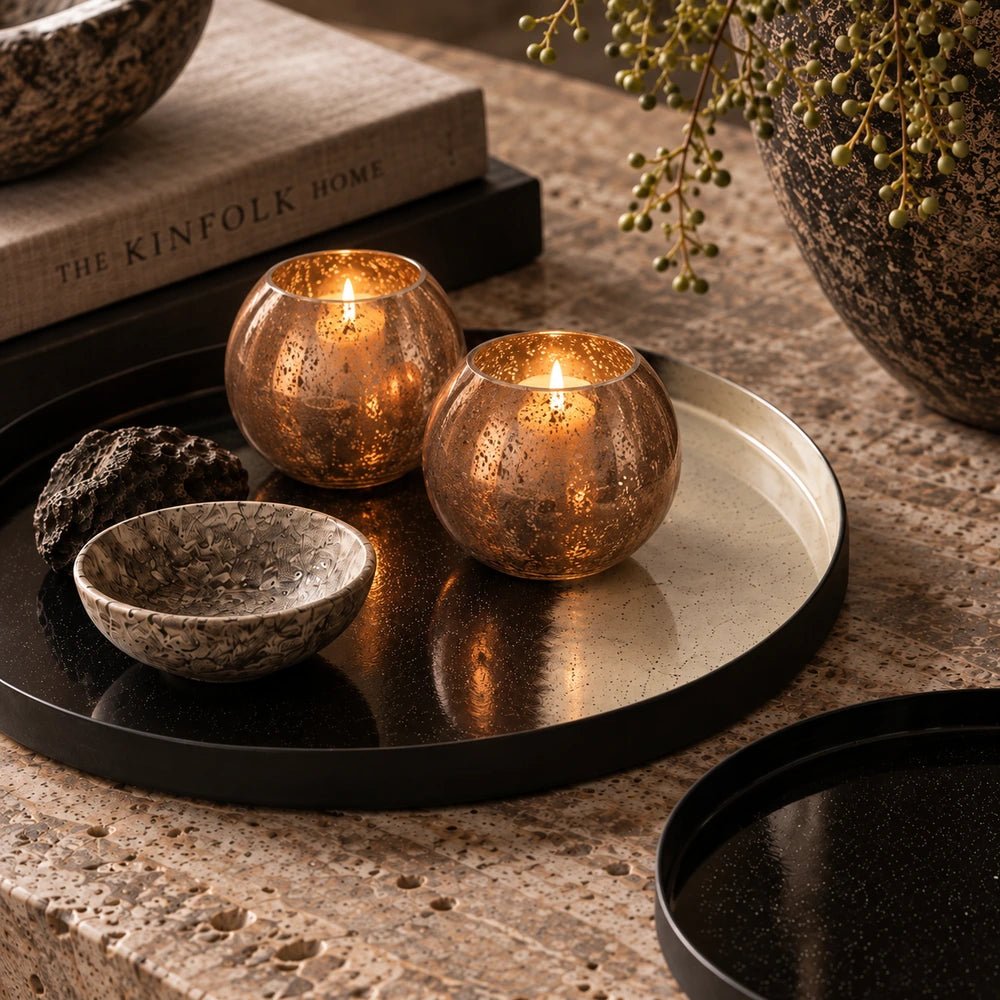 Two Tindra Glass Candle Holders in warm amber textured glass, styled beside stacked books and dark ceramics on a softly lit tabletop.
