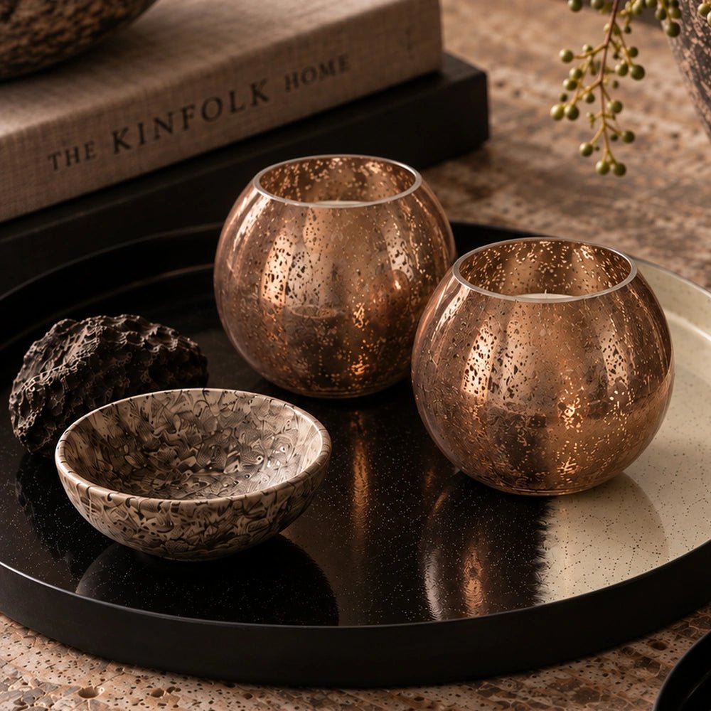 Two Tindra Glass Candle Holders in warm amber textured glass, arranged on a dark tray beside stacked books and a small decorative bowl.