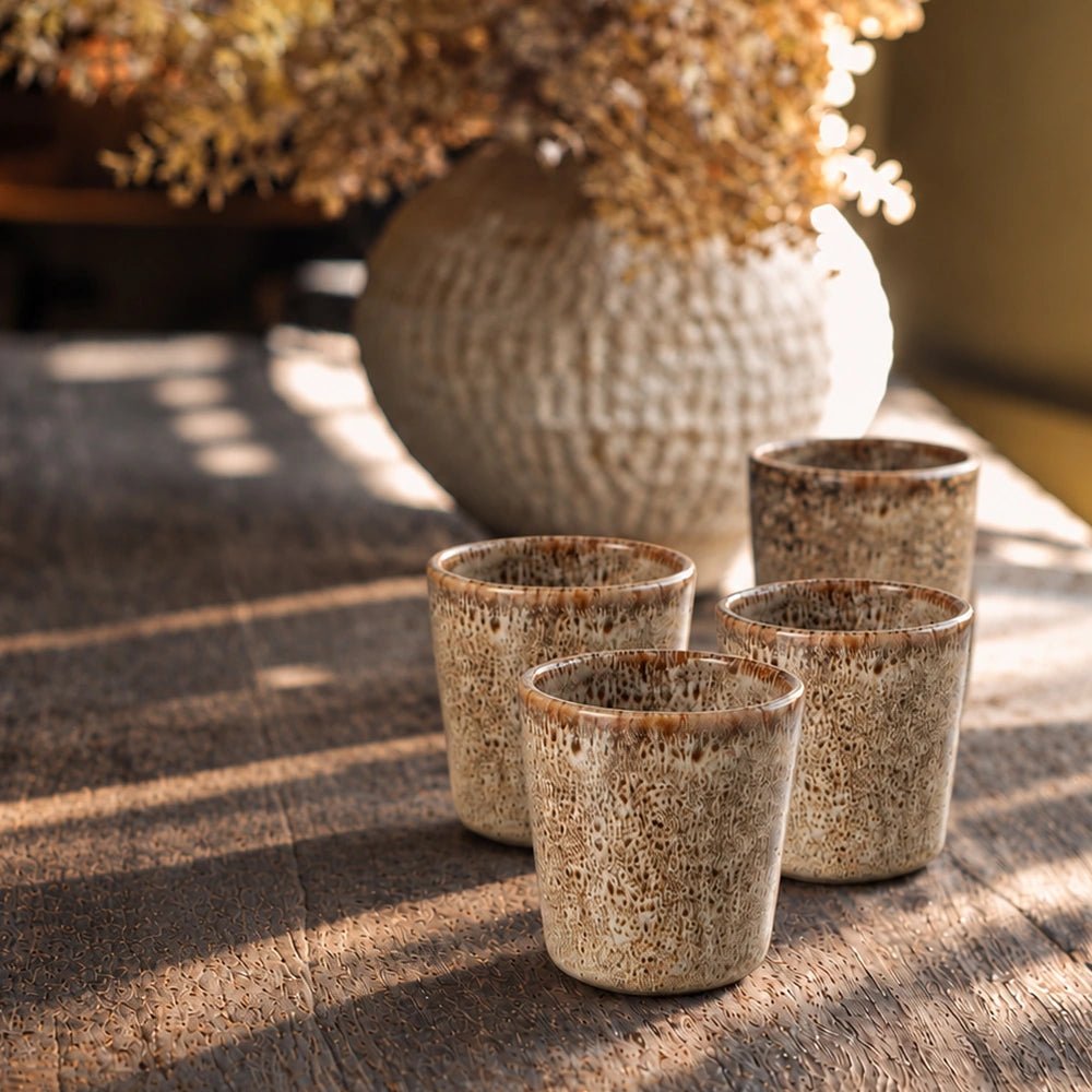 Tisbury Porcelain Cup, Set of 4 represented by one speckled neutral cup on a marble table beside an open book and layered tableware.