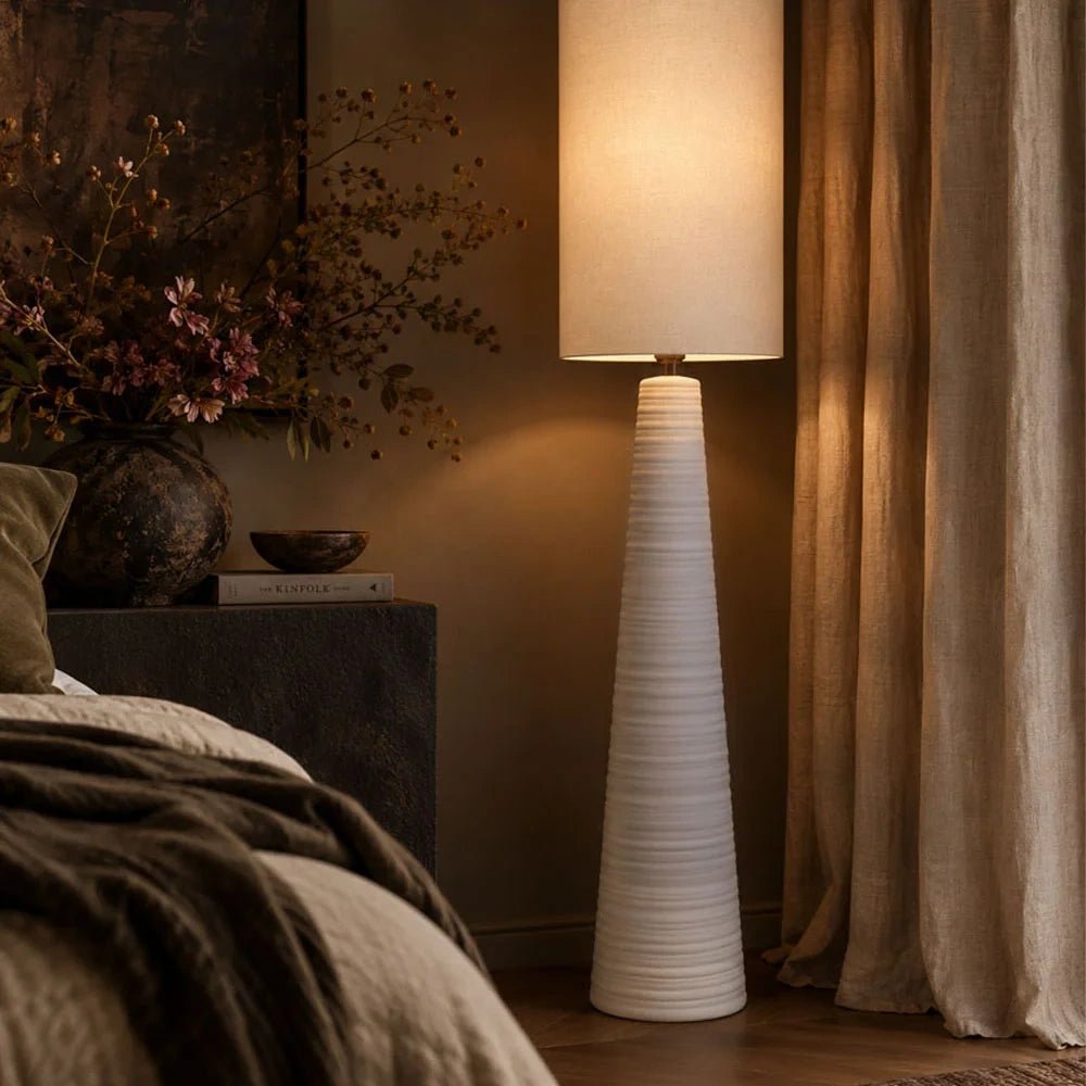 Vanderbilt ceramic floor lamp with a slender tapered white base and tall pale shade, illuminated beside a bed and curtains in a warm neutral bedroom.