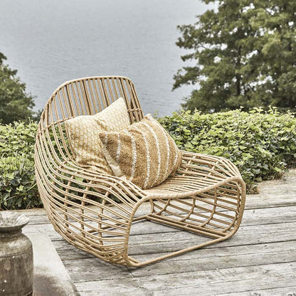 Lifestyle image of our sculptural wicker chair made from PE wicker and iron, styled with a couple of cushions on a wooden deck.
