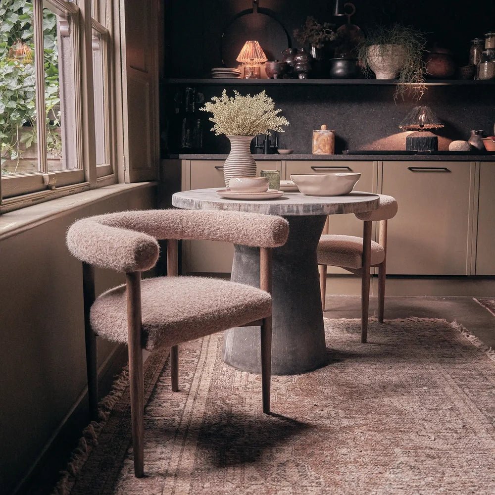 Virgil upholstered dining chairs surround a round pedestal table beside a window, with taupe textured seats, curved arms and slender dark legs on a rug.