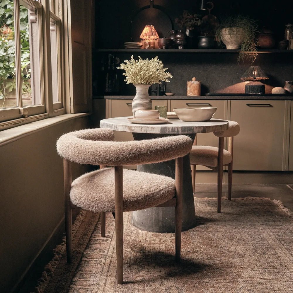 Virgil upholstered dining chair with a curved wraparound back, taupe textured fabric and slim dark legs stands beside a round table in a dining room.