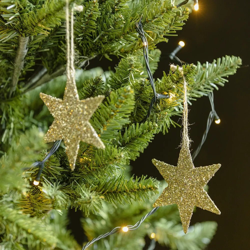Volta glitter star baubles hang at different heights from gold cords among green Christmas tree branches, with warm lights highlighting their flat five-pointed shapes.