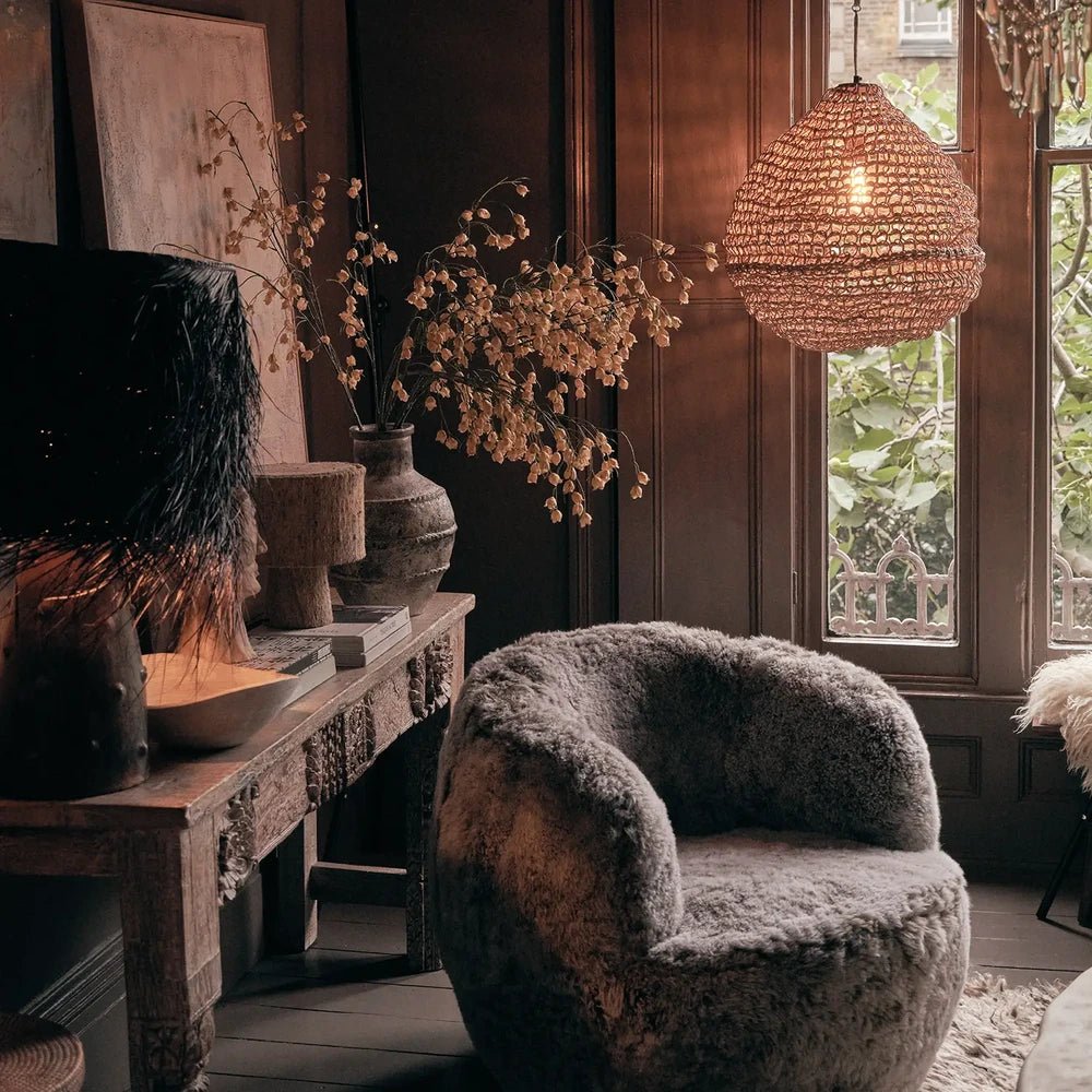 Westcott paper rope pendant hangs beside a tall window in a dark living room, with its warm woven shade above a rounded textured armchair.