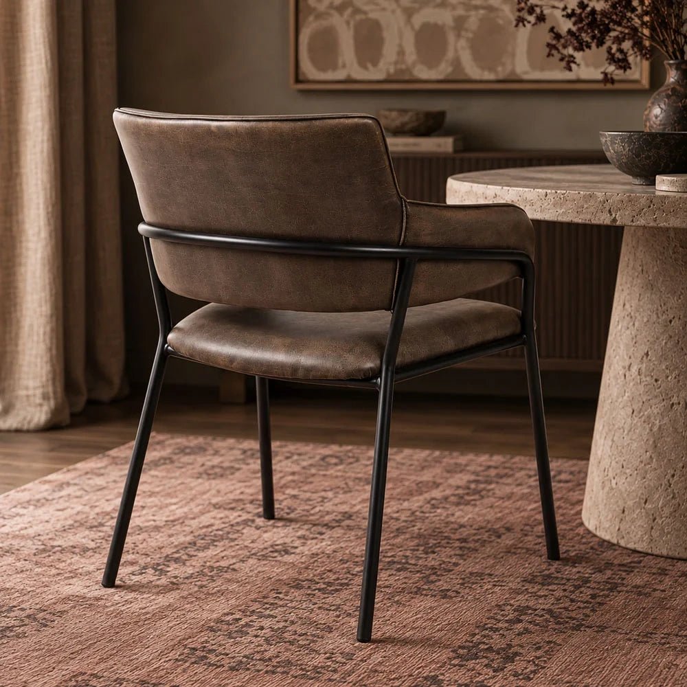 Whitcomb faux leather dining chair viewed from behind beside a round table, showing its dark brown padded back, curved black frame and slender legs.