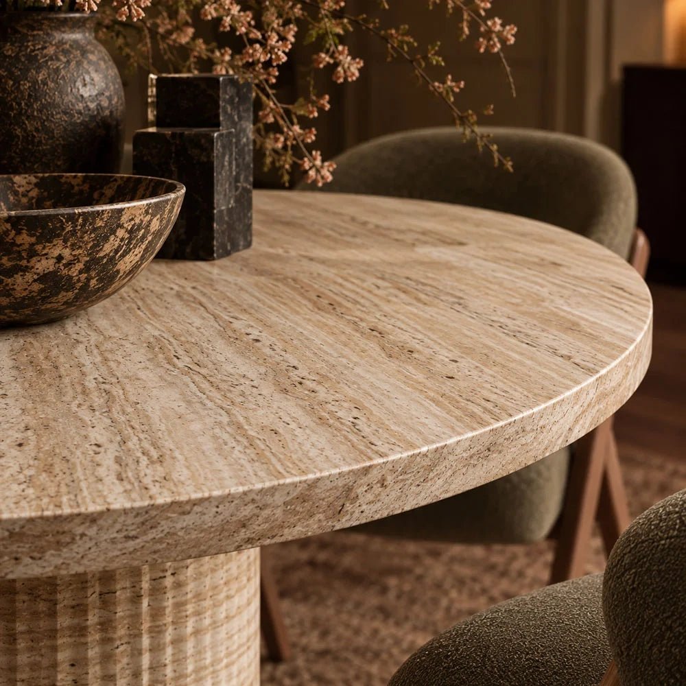 Wilcox faux travertine dining table shown close up, highlighting its round pale patterned top, rounded edge and cylindrical pedestal beneath decorative bowls and a vase.