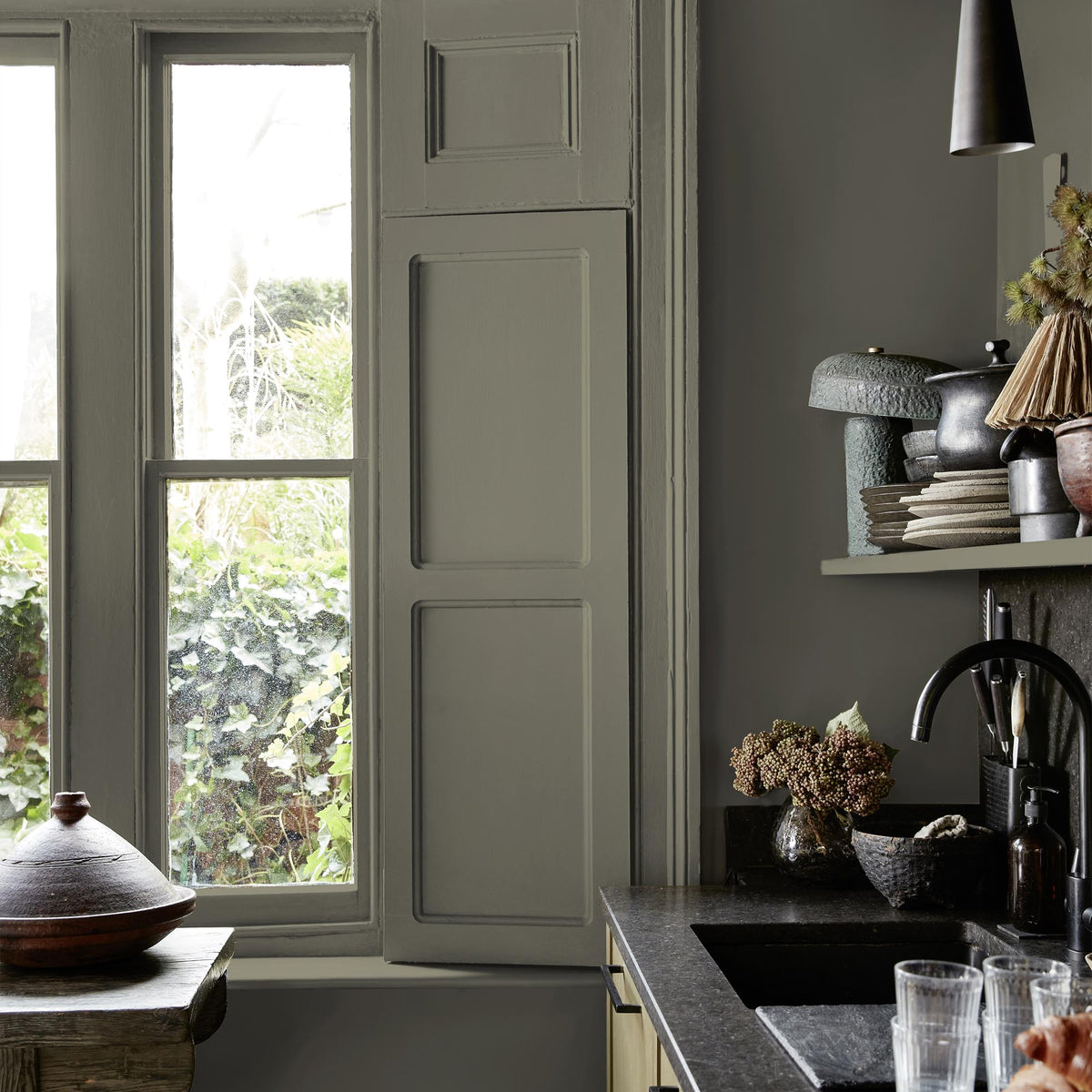 Abigail Ahern Wild Millet paint – a sumptuous light khaki with warm sage green undertones on a kitchen window shutter and wall, beautifully offset by a sleek black countertop